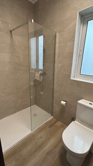 A bathroom with a glass shower enclosure, a toilet, and a window above the toilet.