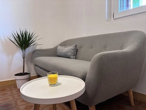 Ein graues Sofa mit einem Kissen, einer kleinen Topfpflanze und einem Glas Orangensaft auf einem weißen Tisch.