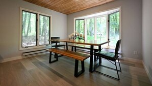 Zona pranzo con tavolo in legno, panche e sedie, ampie finestre con vista sulla foresta e soffitto in legno.