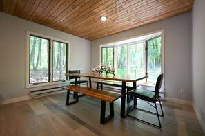 Ein Essbereich mit Holztisch, Bänken und Stühlen, großen Fenstern mit Blick auf einen Wald und einer Holzbalkendecke.