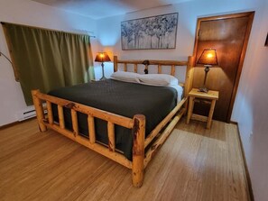 A wooden bed with a green bedspread, two bedside lamps, a wooden nightstand, a green curtain, and a painting on the wall.