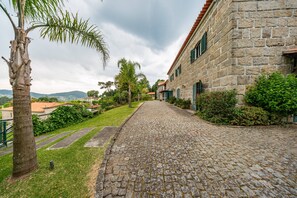 Un sendero adoquinado que lleva a un edificio de piedra con techo de tejas rojas, rodeado de vegetación y una palmera.