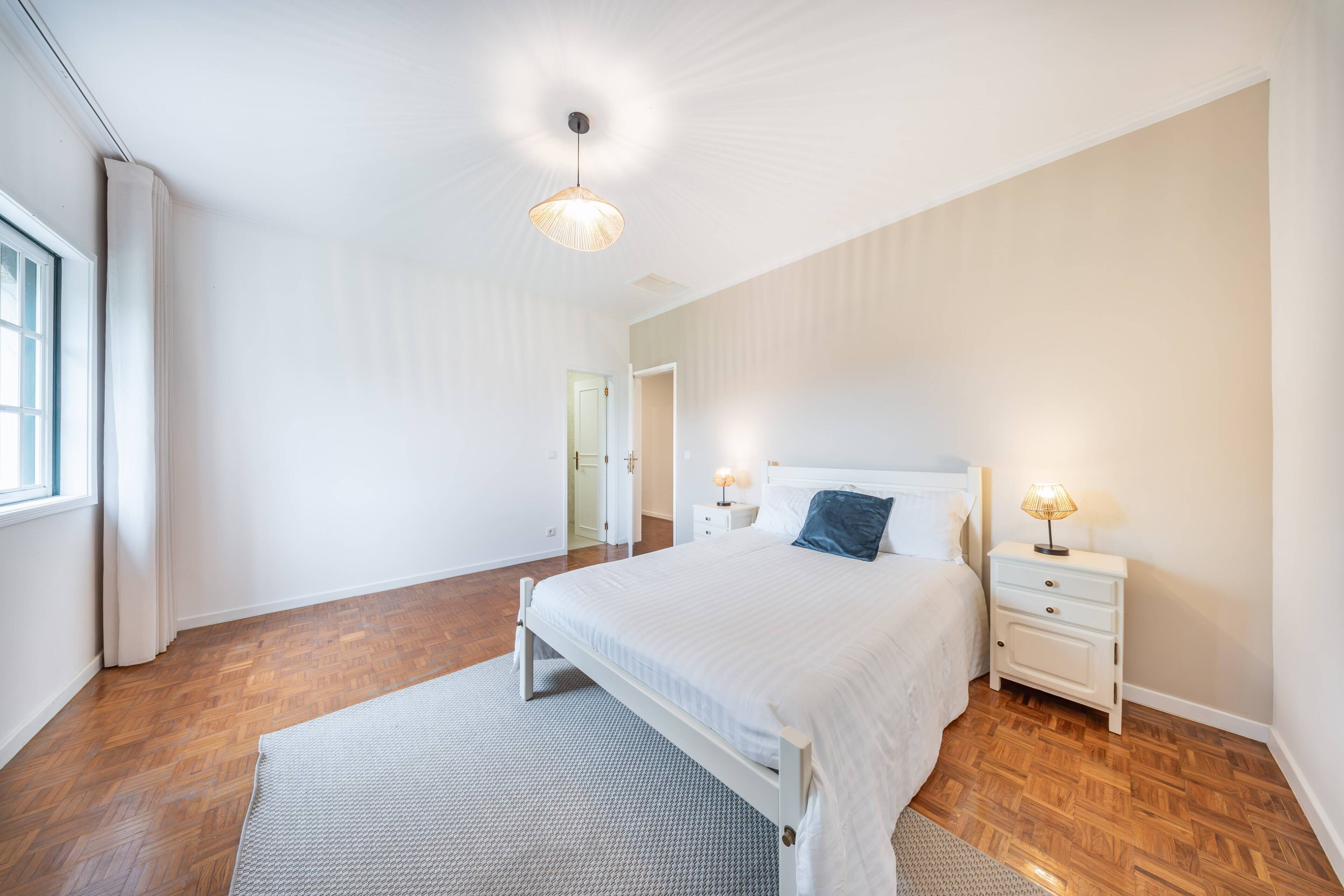 A bedroom with a bed, nightstands, a lamp, a ceiling light, and a door leading to another room.