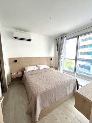 Ein Hotelzimmer mit Bett, Nachttischen, einer Klimaanlage, einem Fenster mit Vorhängen und Blick auf ein Gebäude.