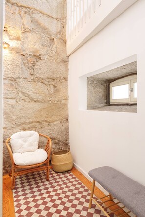 Habitació amb paret de pedra, una cadira de vimet, una banca, un terra de pissos i una petita finestra.