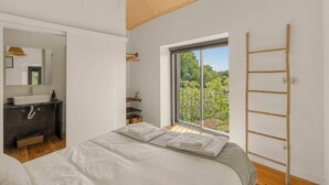 Um quarto com cama, janela com vista para vegetação, banheiro com pia e espelho, e uma escada de madeira encostada na parede.