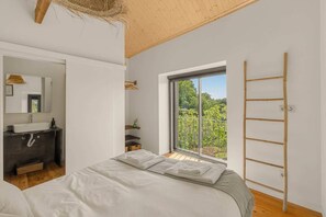 A bedroom with a bed, a window with a view of greenery, a bathroom with a sink and mirror, and a wooden ladder against the wall.
