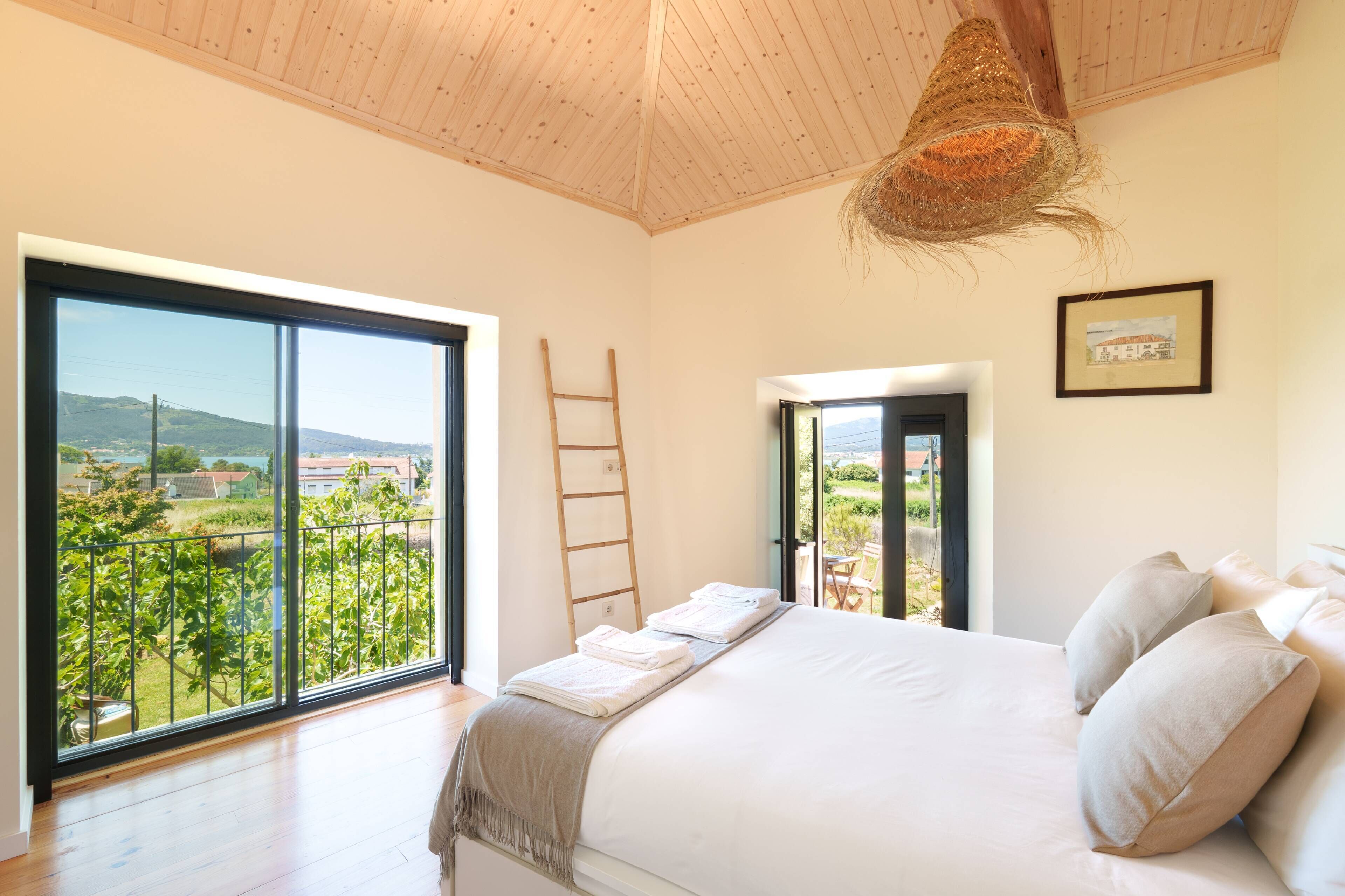A bedroom with a large window, a bed with pillows, a ladder, and a door leading to an outdoor area.