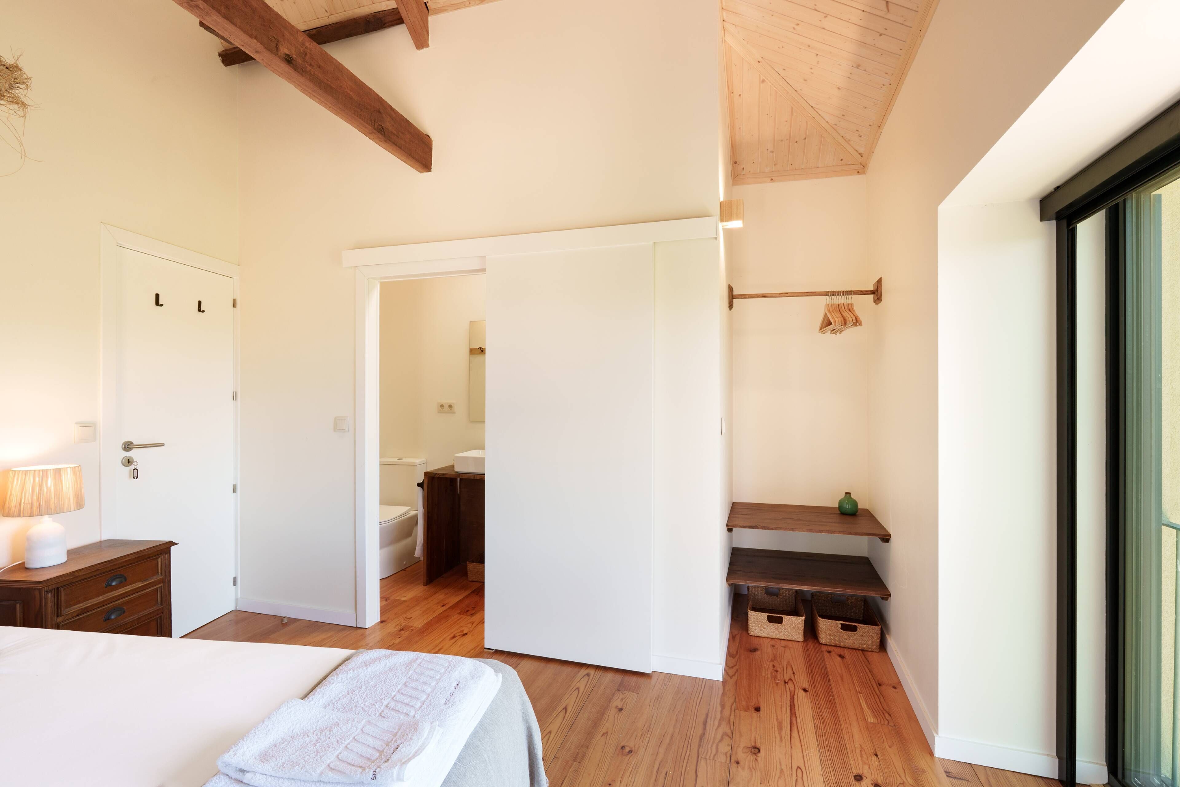 Une chambre à coucher avec un lit, un plancher en bois, une porte, une table de chevet, une lampe, une fenêtre et une vue sur une salle de bain.