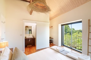 A bedroom with a large bed, a window with a view of greenery, and a bathroom with a mirror and sink. - VIVA Countryside Escape Minho (Caminha)