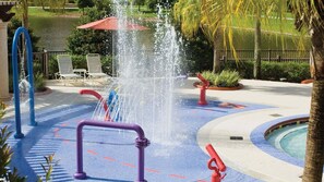 Un área de juegos acuáticos con una fuente central, piscina circundante y sillas de descanso.