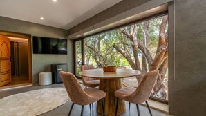 A modern dining area with a round table, four chairs, and a large window with a view of trees. - Olive Tree House - Paderne, Albufeira (Albufeira)