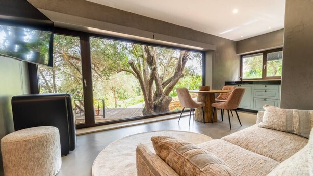 A modern living room with a large window, a round coffee table, a sofa, and a dining area with chairs. - Olive Tree House - Paderne, Albufeira (Albufeira)