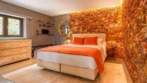 A bedroom with a stone wall, a bed with orange bedding, a dresser, and a mirror. - Olive Tree House - Paderne, Albufeira (Albufeira)
