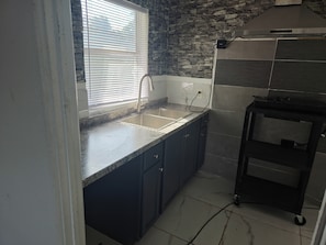 Una cocina con encimera de granito, pileta de acero inoxidable, muebles oscuros, una ventana con persianas y un carrito negro.