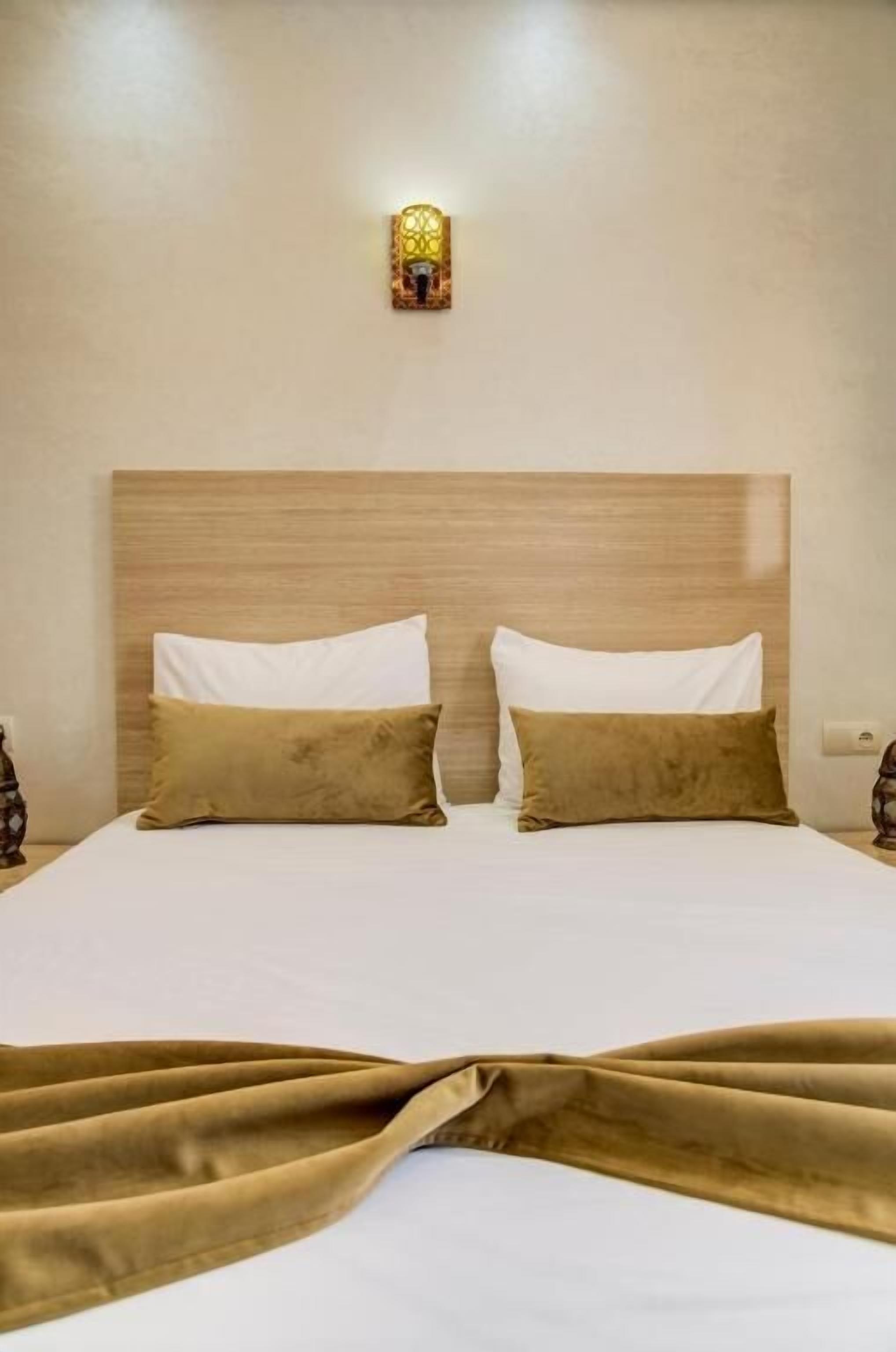 A hotel room with a wooden headboard, two white pillows, a beige blanket, and a wall-mounted light fixture.