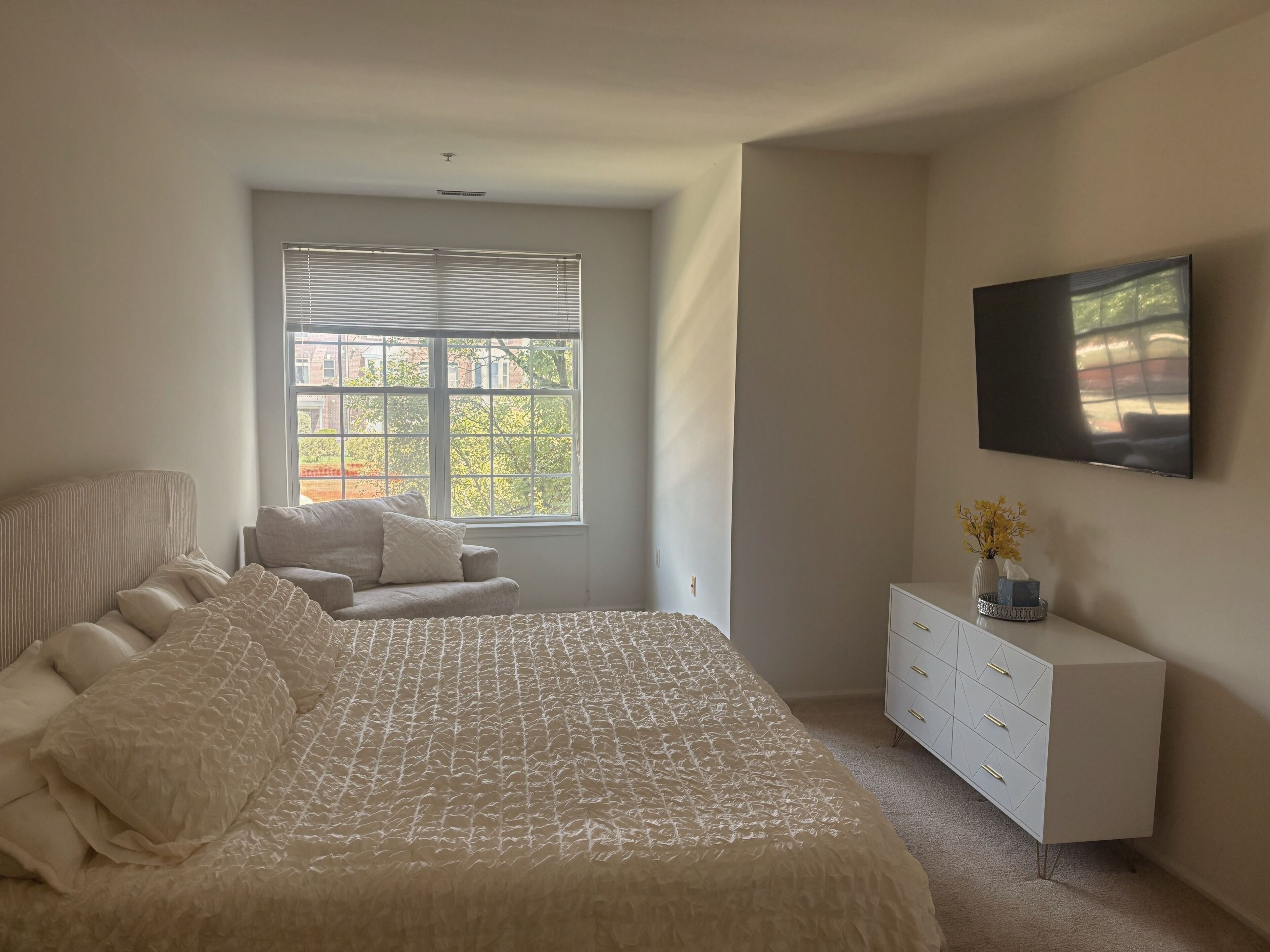 A bedroom with a large bed, a window with blinds, a TV on the wall, and a white dresser.