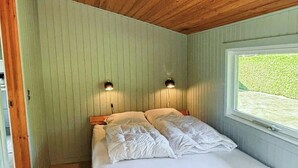 Yhden hengen sänky, jossa on puinen runko, kaksi yöpöytävalaisinta ja ikkuna, josta avautuu näkymä vehreään maisemaan.