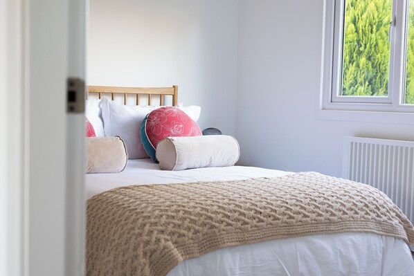 A bedroom with a wooden headboard, white bedding, and a view of greenery through a window.