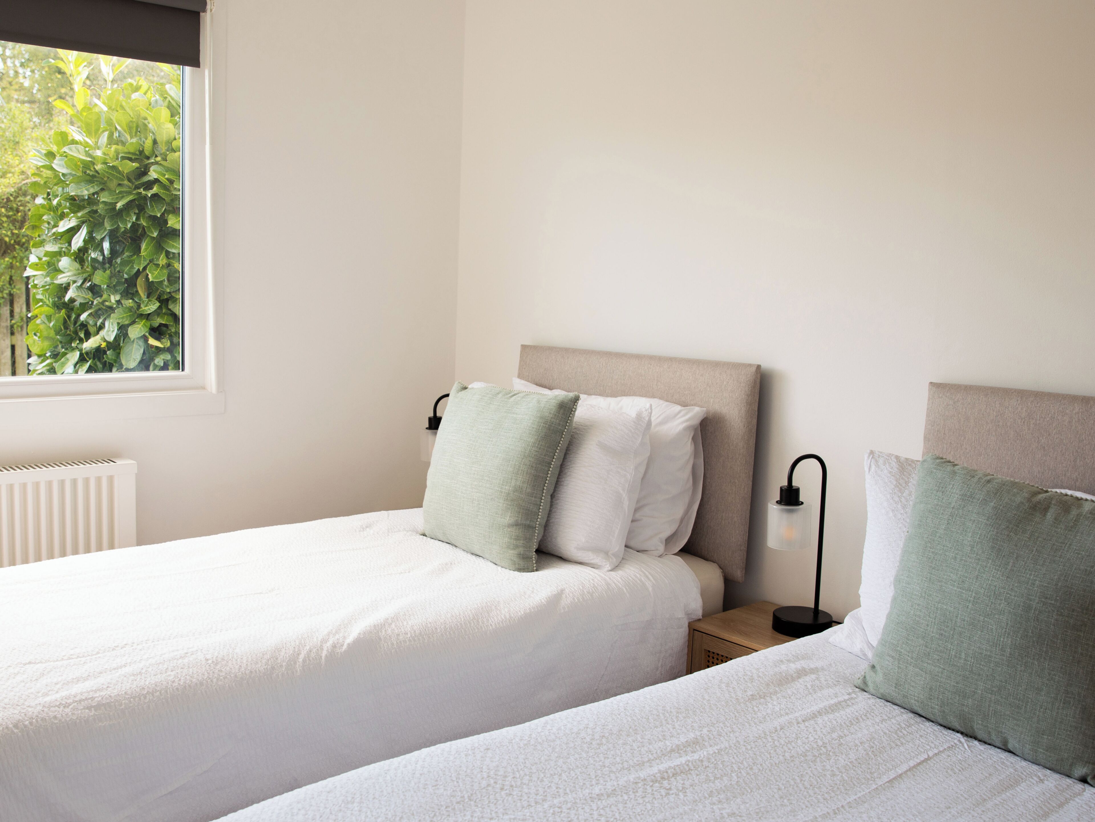 A bedroom with two beds, a window with a view of greenery, a radiator, and a bedside table with a lamp.