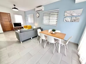 A modern living room with a grey sofa, a wooden dining table with white chairs, a TV, and a ceiling fan. - Charming Villa | Pool & Jacuzzi Near Beaches (Bavaro, Punta Cana)