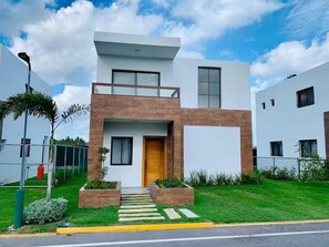 A modern two-story house with a flat roof, a wooden front door, and a small front yard with a pathway leading to the entrance. - Charming Villa | Pool & Jacuzzi Near Beaches (Bavaro, Punta Cana)