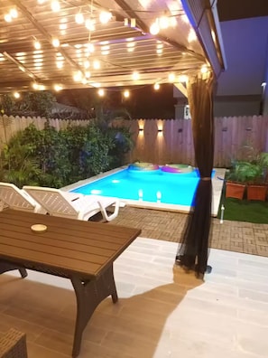 A backyard with a swimming pool, lounge chairs, a table, and string lights. - Charming Villa | Pool & Jacuzzi Near Beaches (Bavaro, Punta Cana)