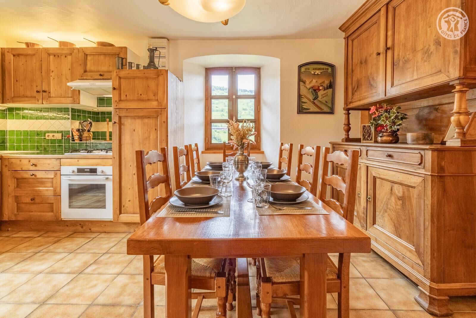 Une table à manger en bois, accompagnée de six chaises, une cuisine avec des meubles en bois et un plan de travail recouvert de carreaux, et une fenêtre donnant sur un paysage verdoyant.