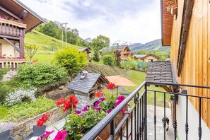 A balcony with a stone wall, a wooden fence, and a view of a village with houses and mountains in the background.