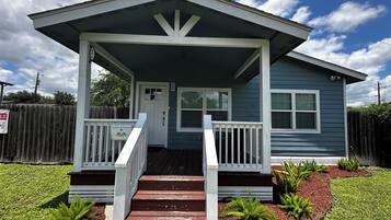 A single-story house with a front porch, a door, and a small front yard with plants.
