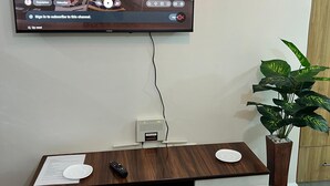 Ein an der Wand befestigter Fernseher zeigt ein Video, darunter eine Holzunterhaltungsecke, eine Fernbedienung, ein kleiner Tisch mit einer Zeitschrift und ein Topfpflanze.