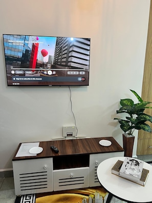 Ein an der Wand befestigter Fernseher zeigt ein Video, darunter eine Holzunterhaltungsecke, eine Fernbedienung, ein kleiner Tisch mit einer Zeitschrift und ein Topfpflanze.
