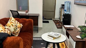Ein kompakter Wohnbereich mit braunem Sofa, einem kleinen runden Tisch, einem Schreibtisch mit Laptop, einem Wasserspender und einem an der Wand befestigten Fernseher.