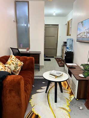 Ein kompakter Wohnbereich mit braunem Sofa, einem kleinen runden Tisch, einem Schreibtisch mit Laptop, einem Wasserspender und einem an der Wand befestigten Fernseher.