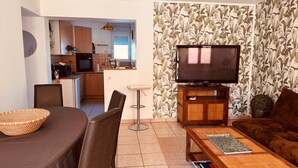 Una sala de estar con televisor, una zona de comedor con mesa y sillas, una cocina al fondo y un papel tapiz estampado.