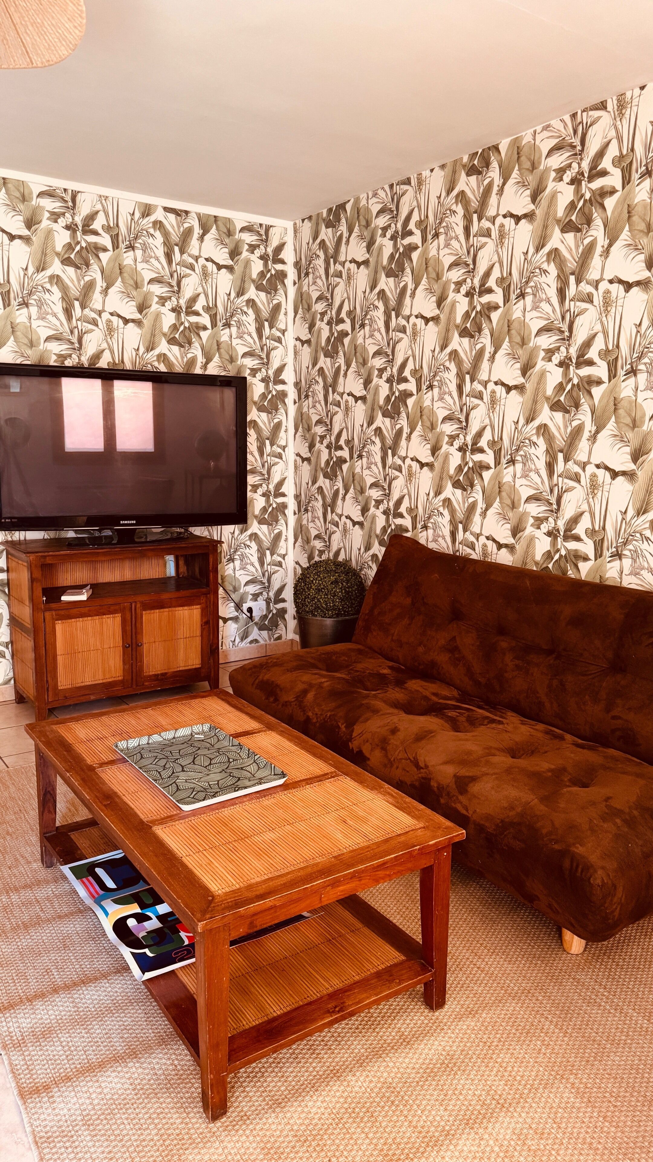 A living room with a television on a wooden cabinet, a brown sofa, a woven coffee table, and a patterned wallpaper.