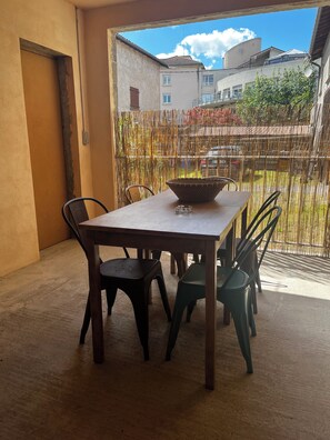 Un coin repas avec une table et des chaises, un panier en osier, et une vue sur les bâtiments et les arbres à travers un paravent en bambou.