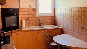 Una cocina compacta con horno integrado, ventana con persianas, un lavamanos y zona de comedor con mesa y sillas.