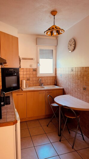 Une cuisine compacte dotée d’un four encastré, d’une fenêtre avec stores, d’un évier et d’un coin repas avec une table et des chaises.