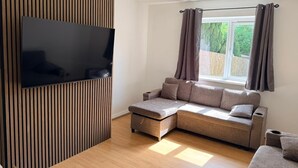 A modern living room with a sectional sofa, a flat-screen TV mounted on a wooden panel, a window with curtains, and a ceiling light. -  Semi-Detached w/Pool Table & Garden (Cardiff)