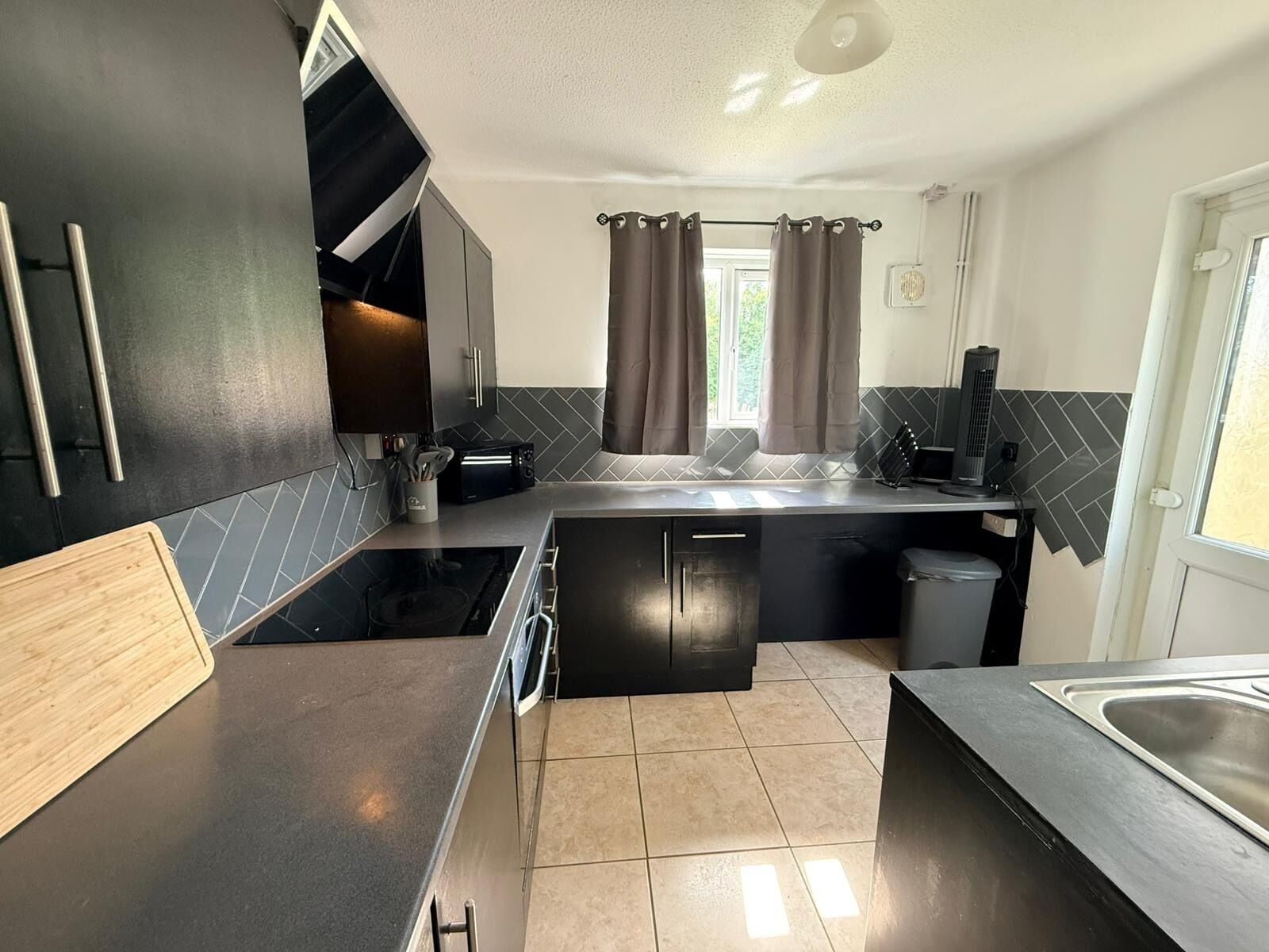 A compact kitchen with a stove, microwave, and sink, a small window with curtains, and a door with a light fixture..  Semi-Detached w/Pool Table & Garden