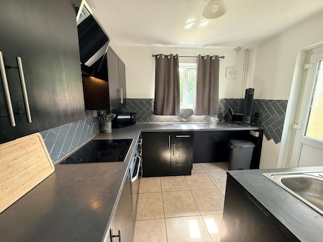 A compact kitchen with a stove, microwave, and sink, a small window with curtains, and a door with a light fixture.. Semi-Detached w/Pool Table & Garden