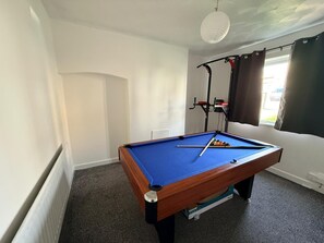 Una mesa de billar en una habitación con paredes blancas, un radiador y una ventana con cortinas.