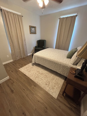 Un dormitorio con cama, una silla, una mesita de noche, una lámpara, un cuadro enmarcado en la pared y un ventilador de techo.