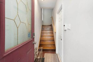 A narrow hallway with a wooden staircase, a red door with a geometric pattern, and a white wall with a thermostat. - Modern Comfort in Olde Walkerville (Windsor)