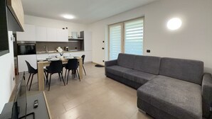 Eine moderne Küche mit weißen Schränken, einem Esstisch mit Stühlen, einem grauen Sofa und einem großen Fenster mit Jalousien.