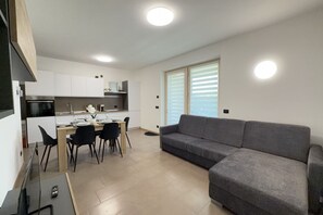 A modern kitchen with white cabinets, a dining table with chairs, a gray sofa, and a large window with blinds. - 20 min from Gardaland and lake with swimming pool (Valeggio sul Mincio)