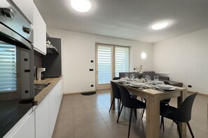 A modern kitchen with a dining area, featuring a wooden table set for four, a built-in oven, and a window with blinds. - 20 min from Gardaland and lake with swimming pool (Valeggio sul Mincio)