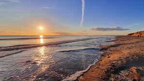 Una spiaggia con il sole che tramonta sull'oceano, le onde che lambiscono dolcemente la riva e un cielo sereno.