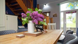 Un tavolo di legno con un vaso bianco pieno di fiori viola, un cartello di benvenuto e una pila di biglietti.